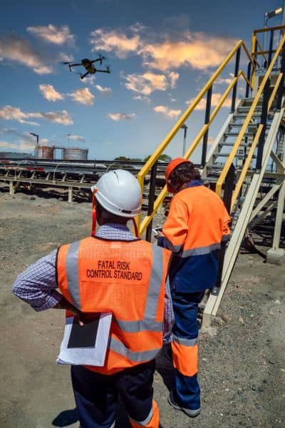 Two surveyors wearing safety vests and helmets monitor a drone surveying an industrial site, highlighting risk management in drone surveying.
